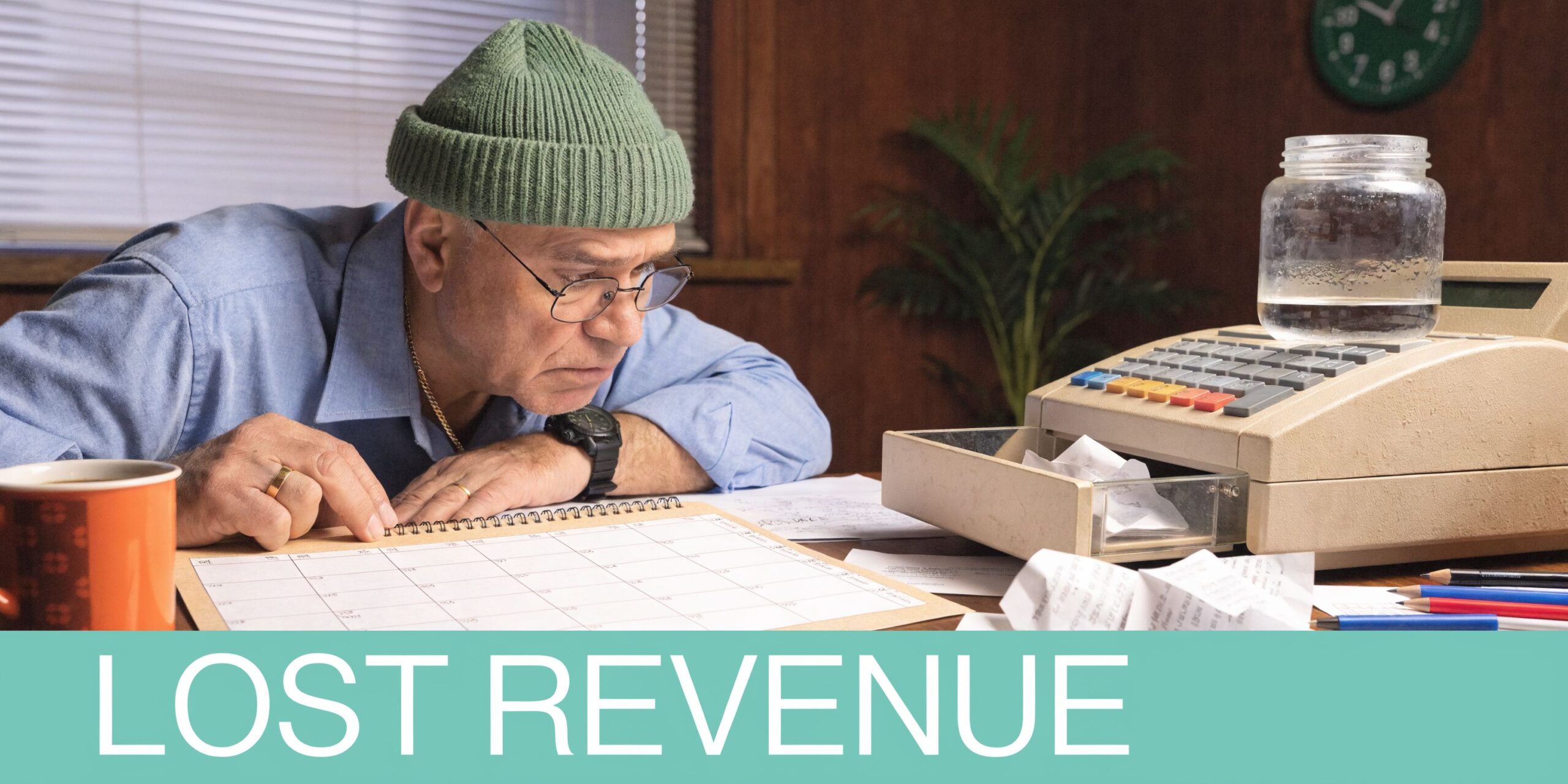 A middle-aged man wearing a beanie and glasses studies a calendar while looking at a cash register.